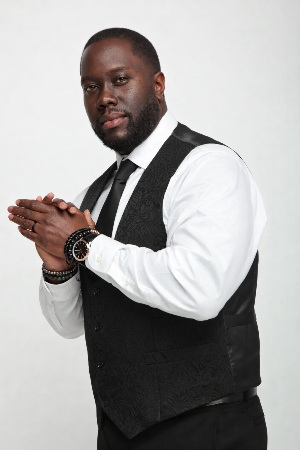 A man dressed in a white shirt, black vest, and black tie, standing with his hands clasped, wearing a watch and bracelets, looking confidently at the camera against a plain background.