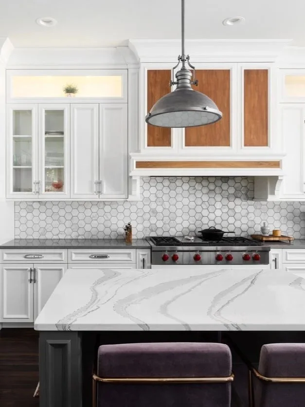 Modern kitchen with white cabinets, a gray and white marble countertop island, hexagonal white backsplash, and a stainless steel industrial pendant light overhead.