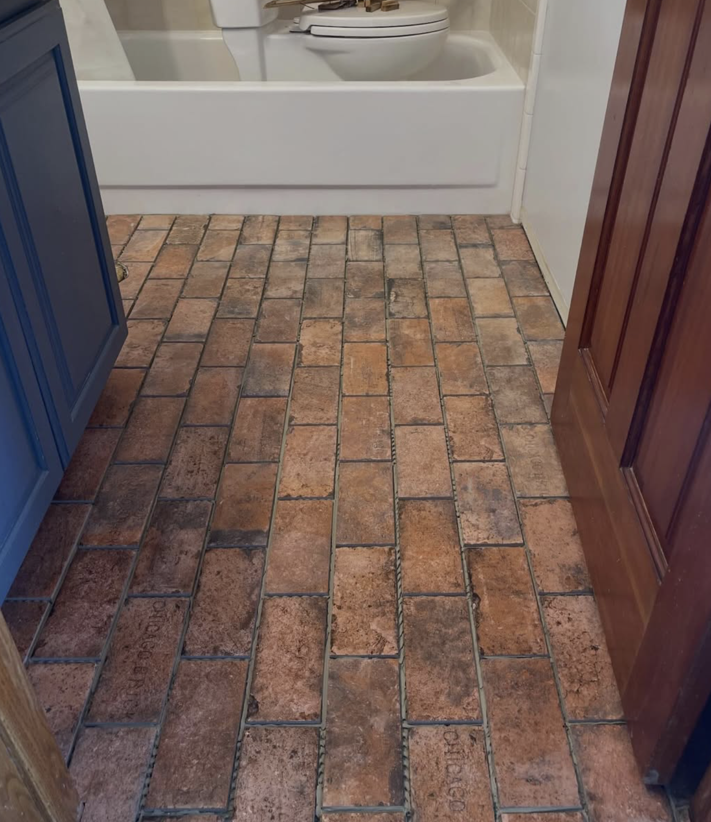View of a bathroom floor with brownish-orange brick tiles, a white bathtub, wooden and blue cabinets.