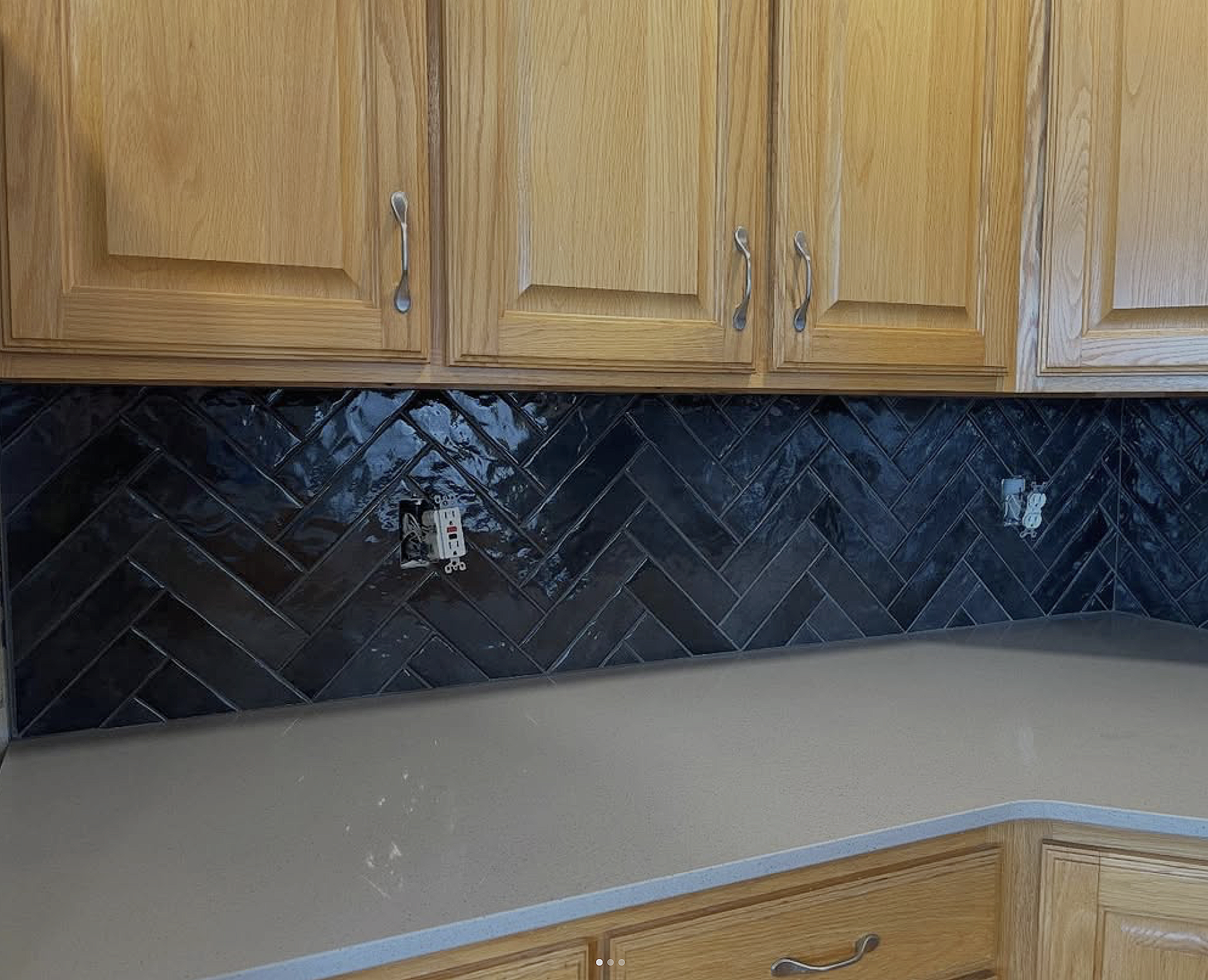 Kitchen wall with dark glossy herringbone tile backsplash, wooden cabinets, and open electrical outlets.