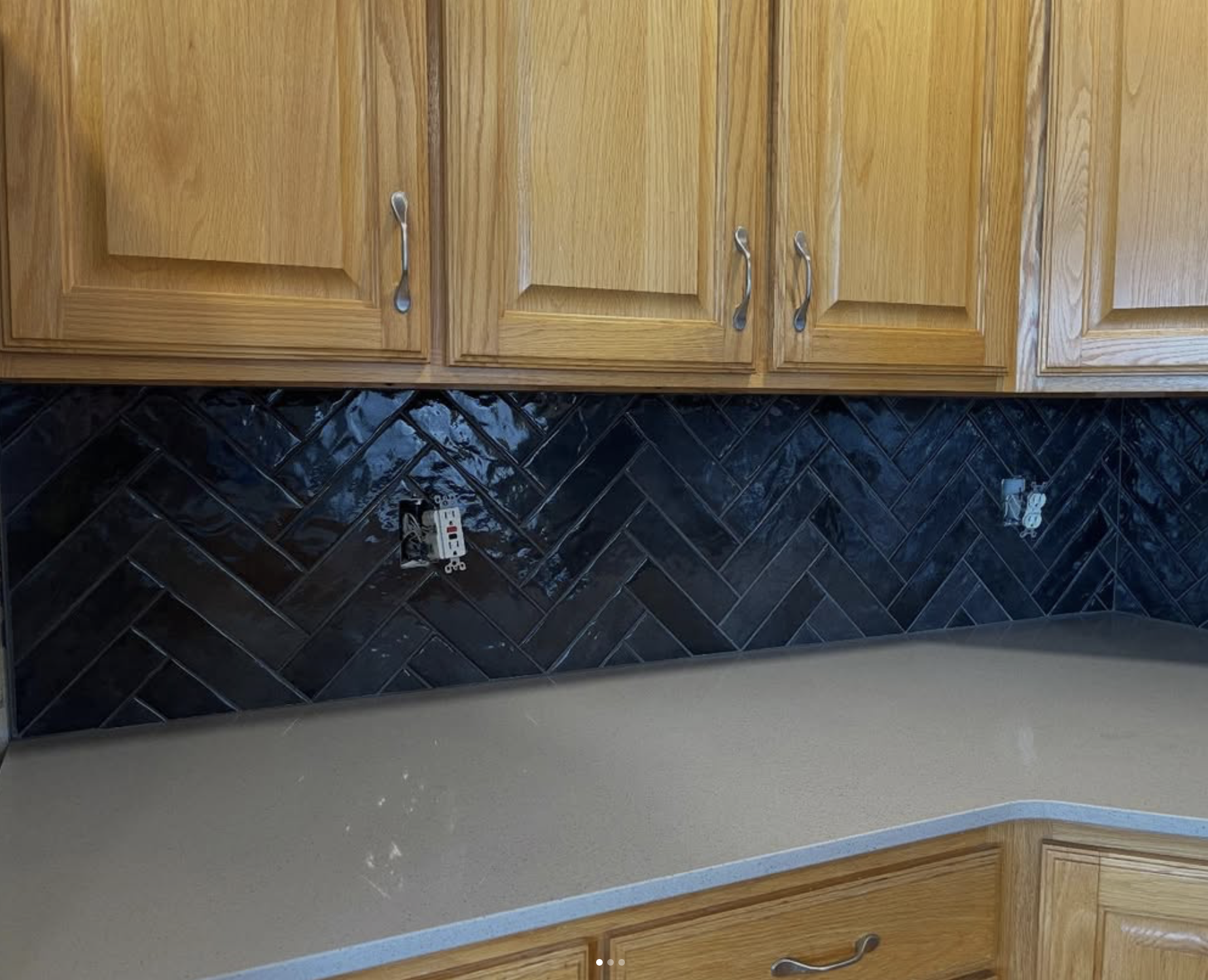Kitchen with wooden cabinets, black backsplash tiles, and a beige countertop. There are two electrical outlets on the backsplash, one with a switch and socket, one unplugged.