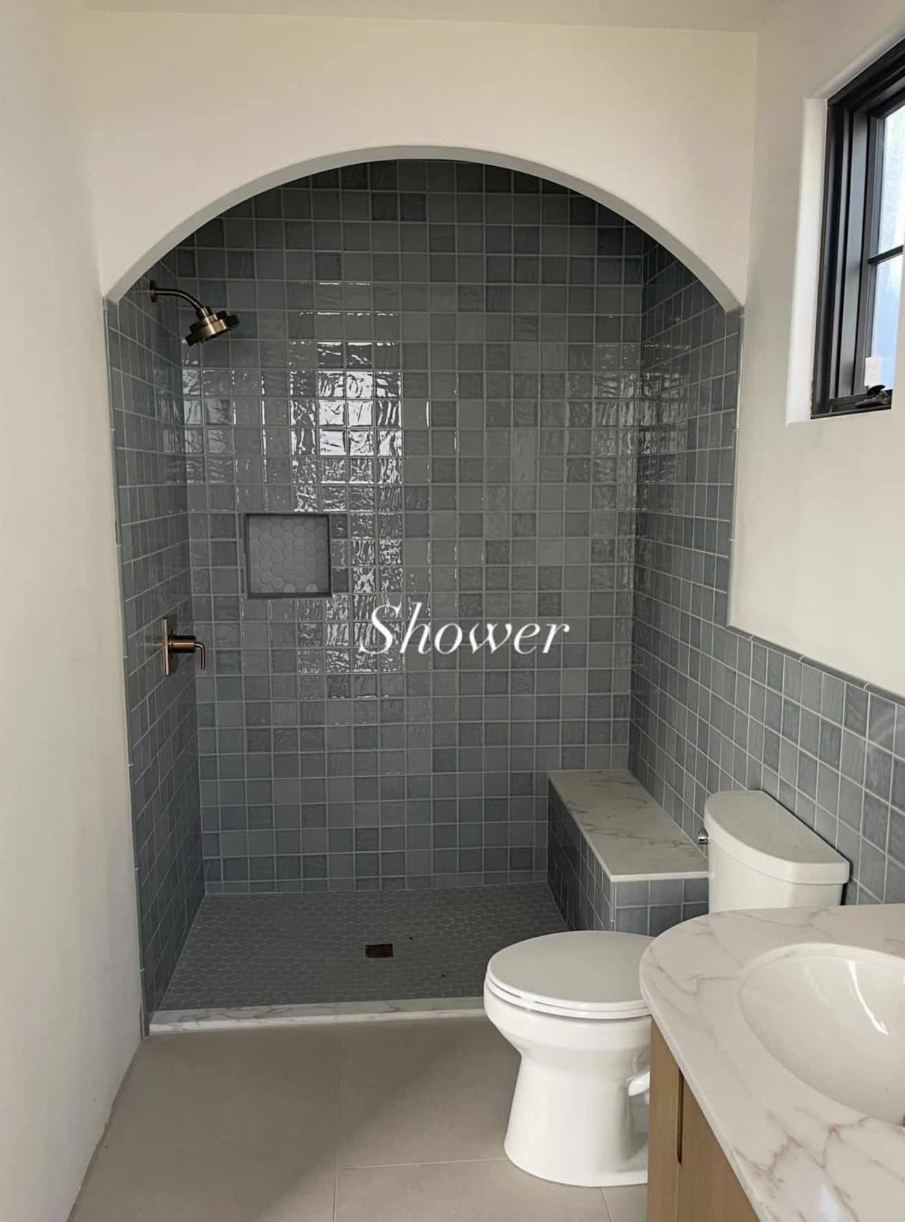 Bathroom with a walk-in shower featuring gray tiles, a small niche, a window, a toilet, and a marble countertop with a sink.