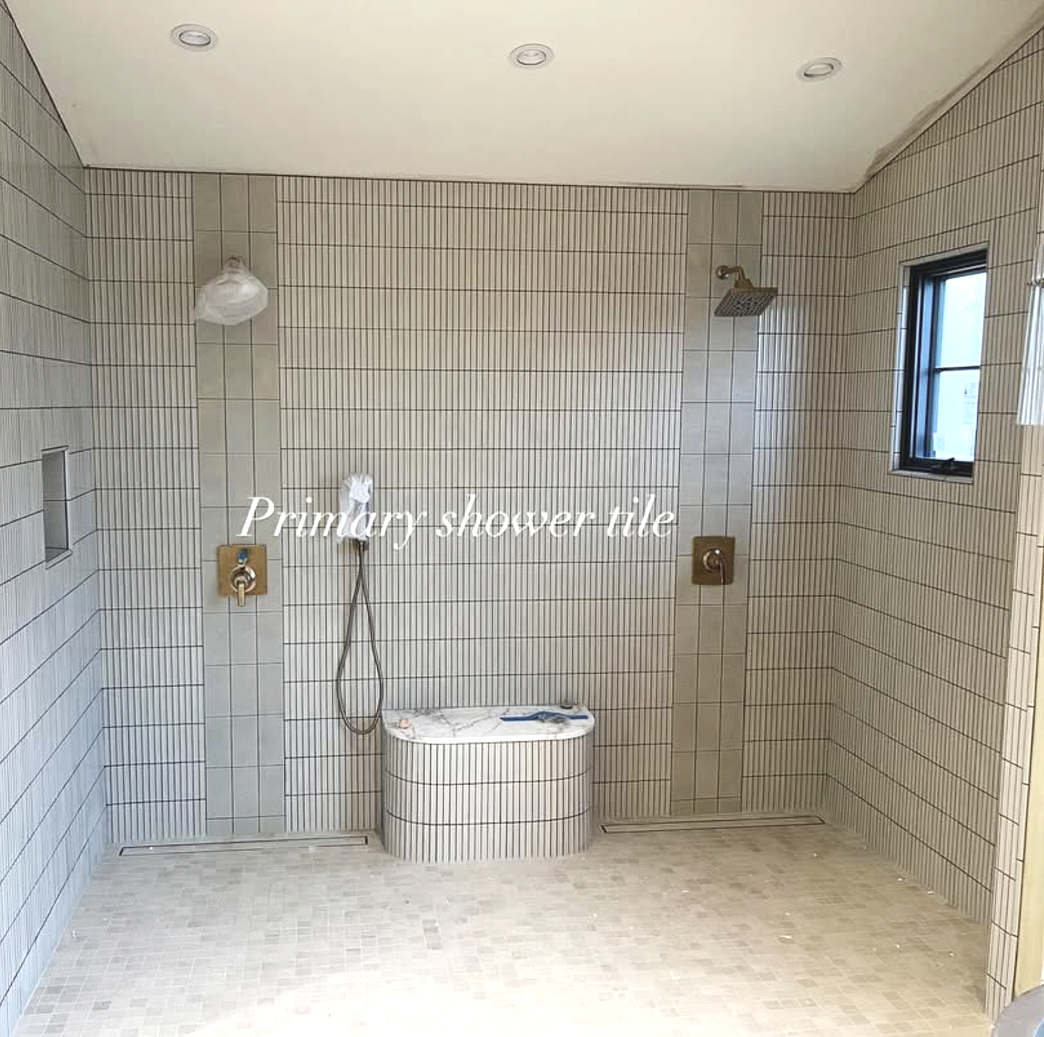 A spacious shower area with beige tiles featuring vertical black stripe accents. The shower has two brass fixtures, one with a handheld showerhead and one overhead rain shower. There is a small window on the right wall and a built-in niche on the left wall. The ceiling has recessed lighting, and there is a small bench or platform in the center. The floor and wall tiles are coordinated with a subtle mosaic pattern.
