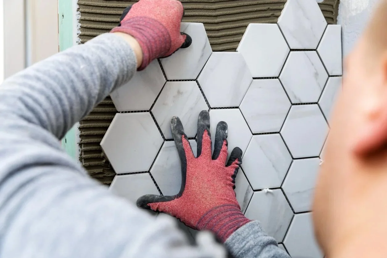 A person installing white hexagonal tiles on a wall, wearing red and black gloves and a gray long sleeve shirt.