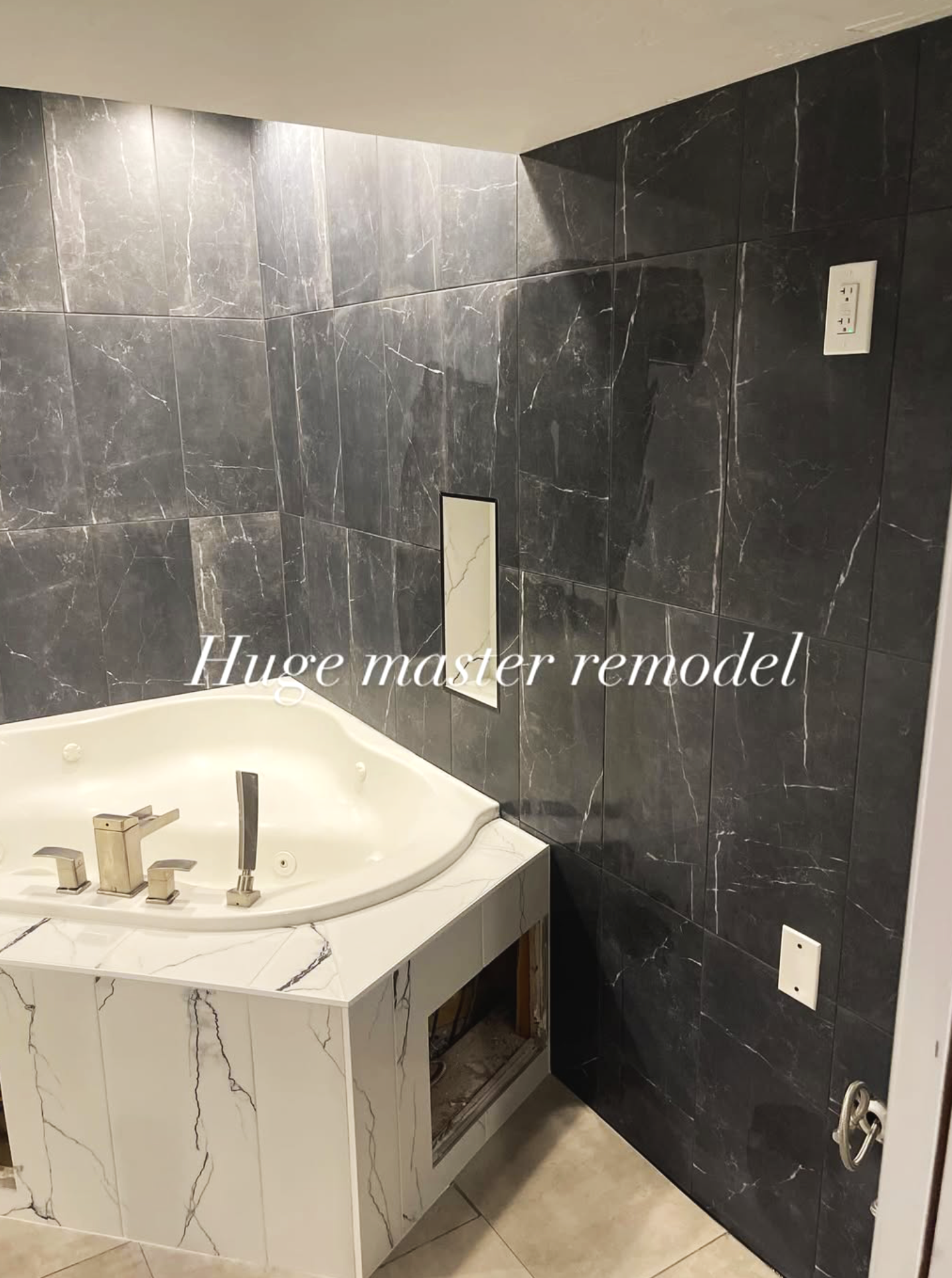 Bathroom with black marble tiles on the wall, a white bathtub with a marble-like facade, and a faucet with three control knobs. There is an electrical outlet and some wiring visible, suggesting ongoing renovation.