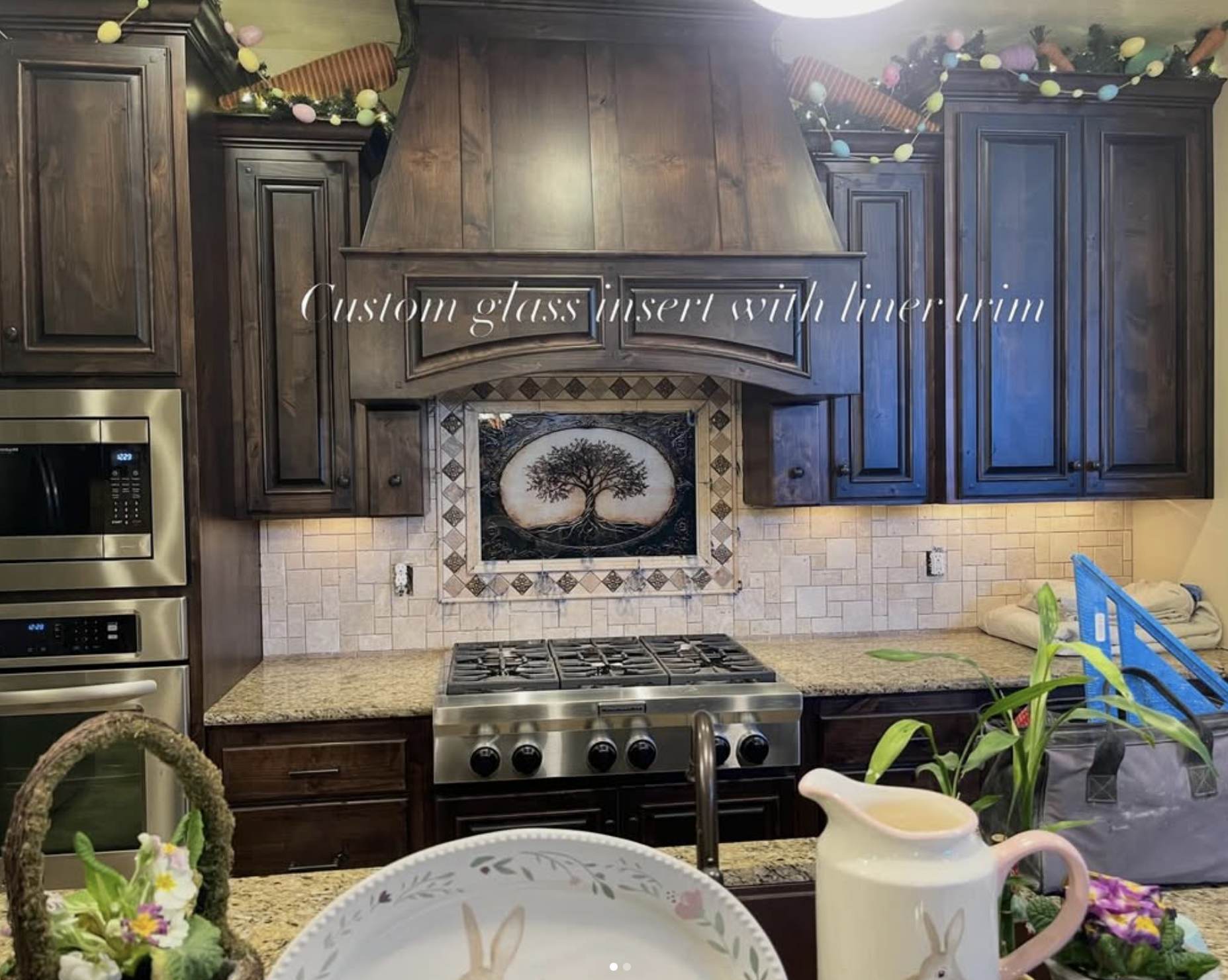 Kitchen with dark wooden cabinets, a granite countertop, and a beige tile backsplash. There is a stove with a decorative tile featuring a tree in the backsplash, and decorated garlands with pastel-colored eggs and ornaments on top of the cabinets. The counter has a white pitcher, some green plants, and a basket with flowers.