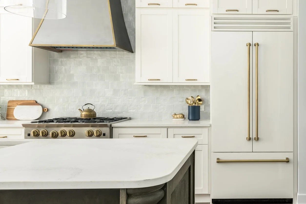 Modern white kitchen with gold hardware, a stovetop with a kettle, microwave, cutting boards, and decorative objects on the counter.