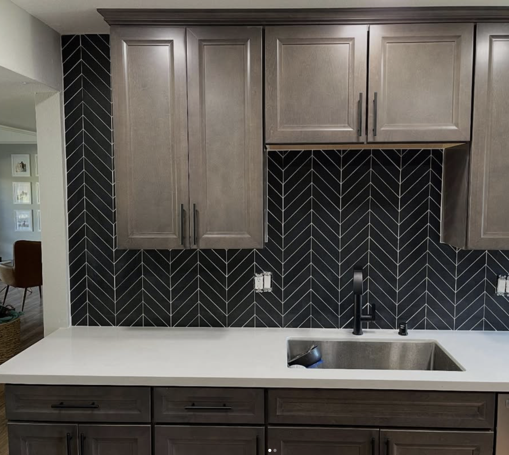 Kitchen countertop with black herringbone tile backsplash, gray wooden cabinets, and a stainless steel sink with a black faucet.