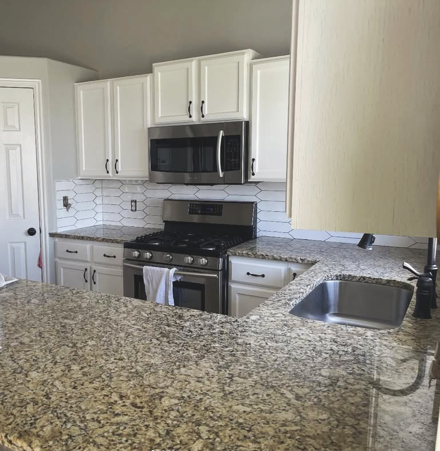 A modern kitchen with granite countertops, white cabinets, and stainless steel appliances including a microwave and oven. There are hexagonal tile backsplashes behind the stove.