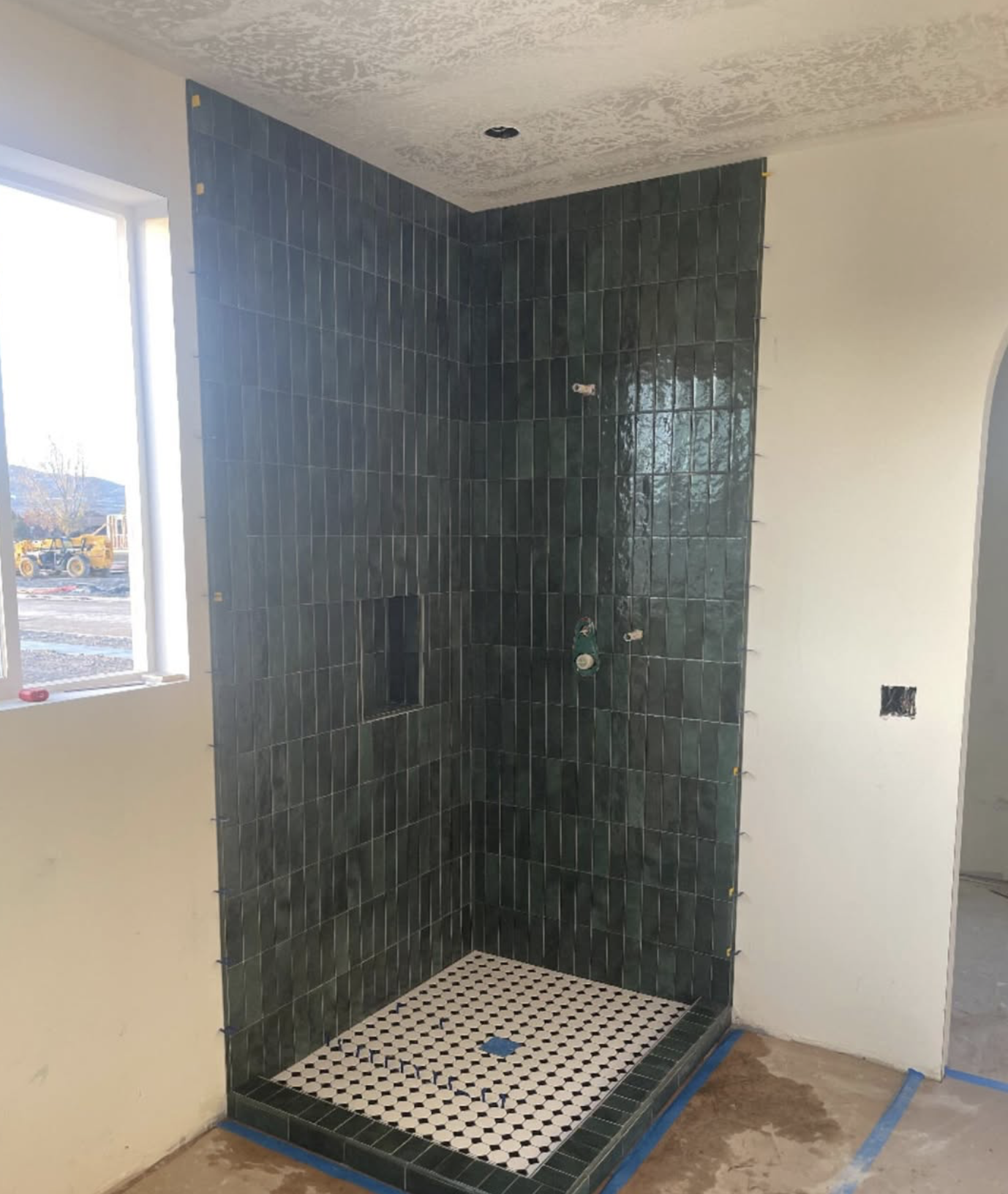 Bathroom under construction with a corner shower area tiled in dark green tiles, a small niche in the shower wall, and a white and black patterned tile floor. An unfinished wall with an electrical outlet and a window nearby.