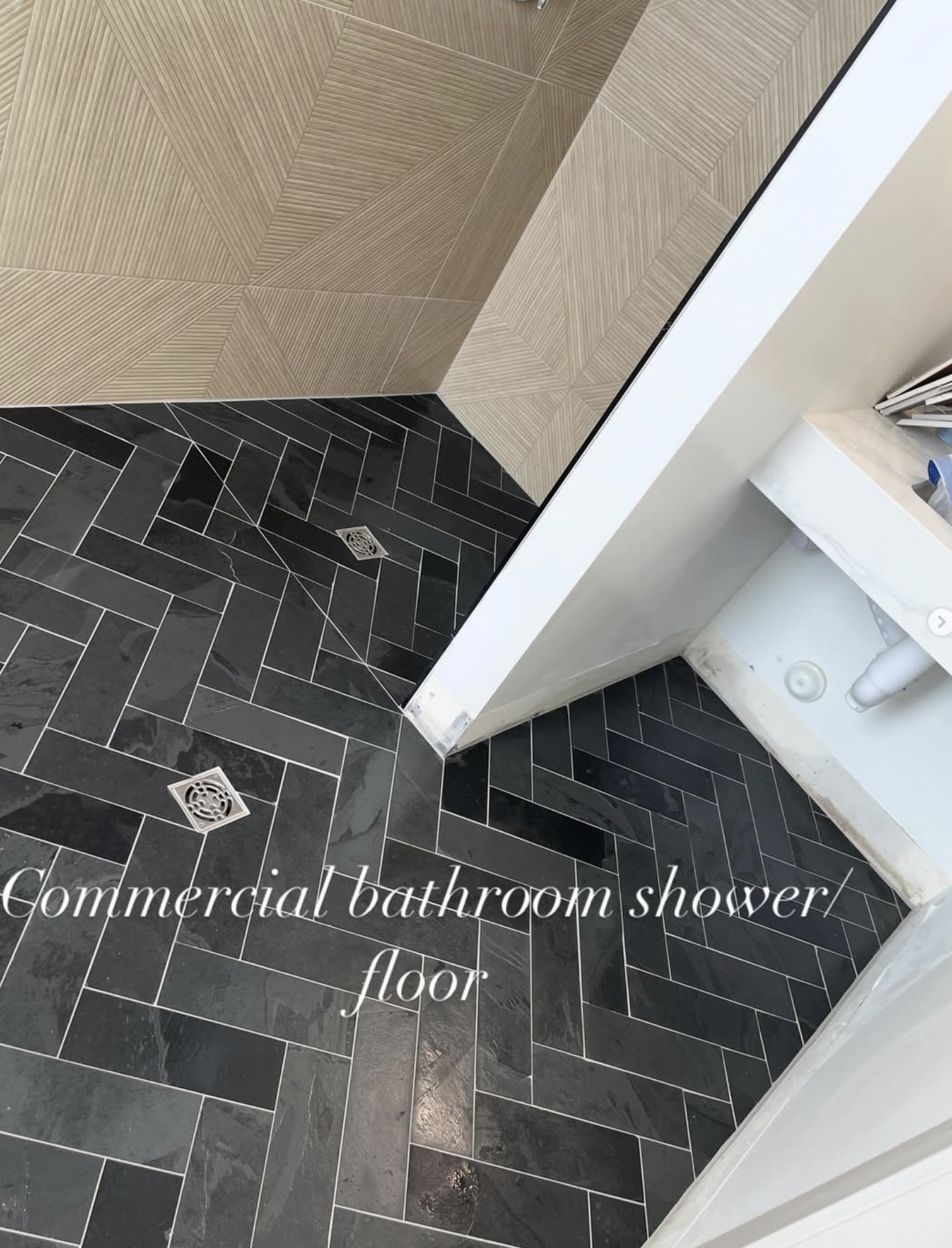 Black herringbone patterned bathroom shower floor with a drain, adjacent to beige wooden textured wall tiles, with part of a white sink and plumbing visible on the right side.