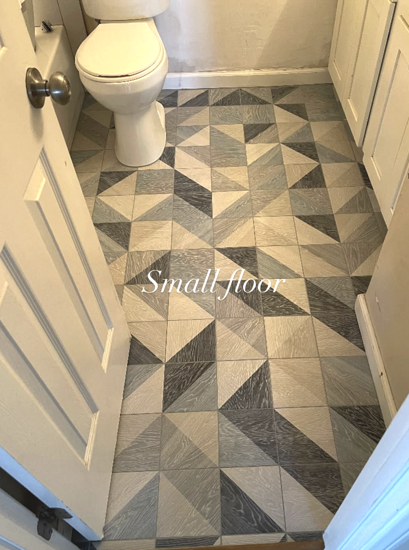A small bathroom with patterned gray and beige tile floor, a white toilet, and white cabinets.