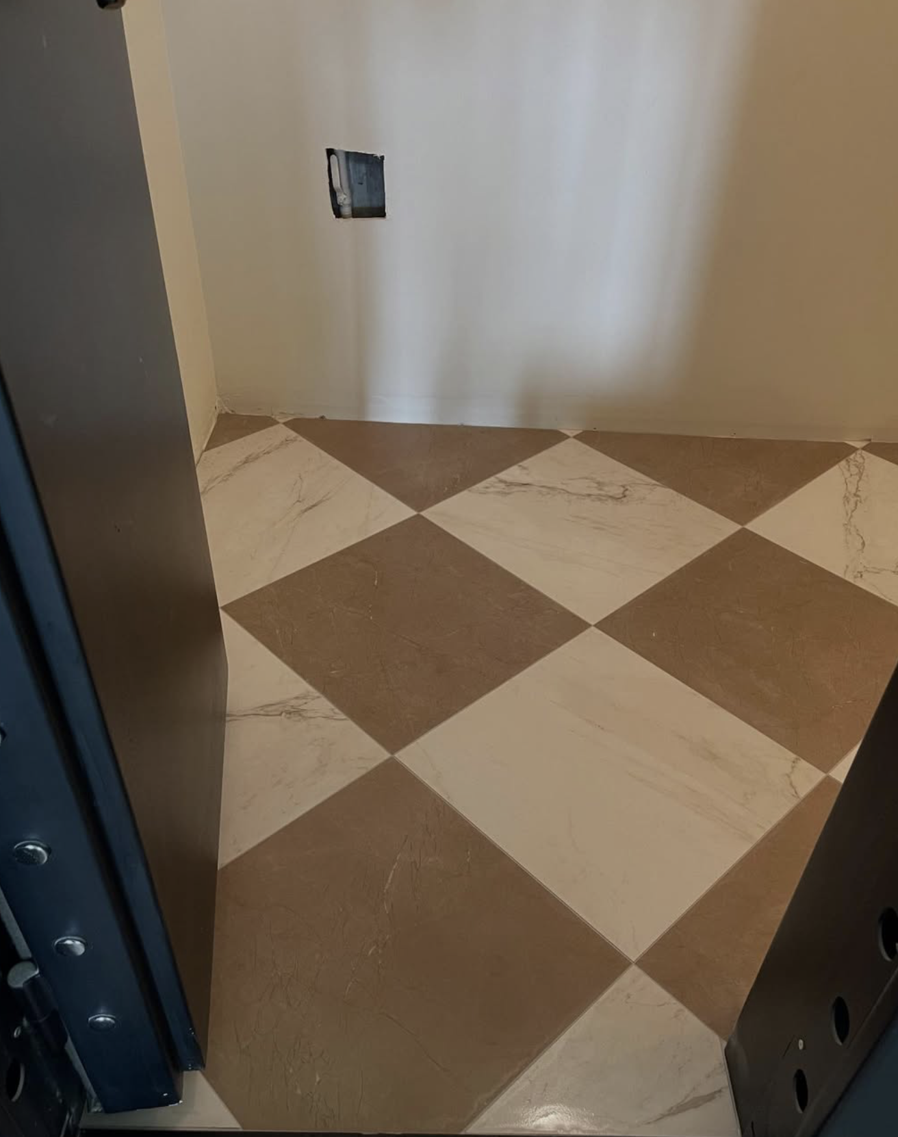 Empty room with a checkered floor in shades of brown and beige, and an open wall outlet on the beige wall.