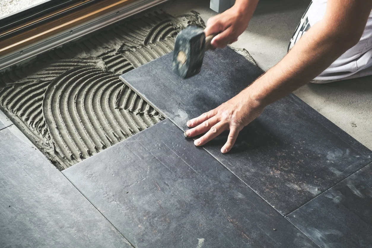 Person installing dark gray tile flooring using a hammer and adhesive on a concrete surface.