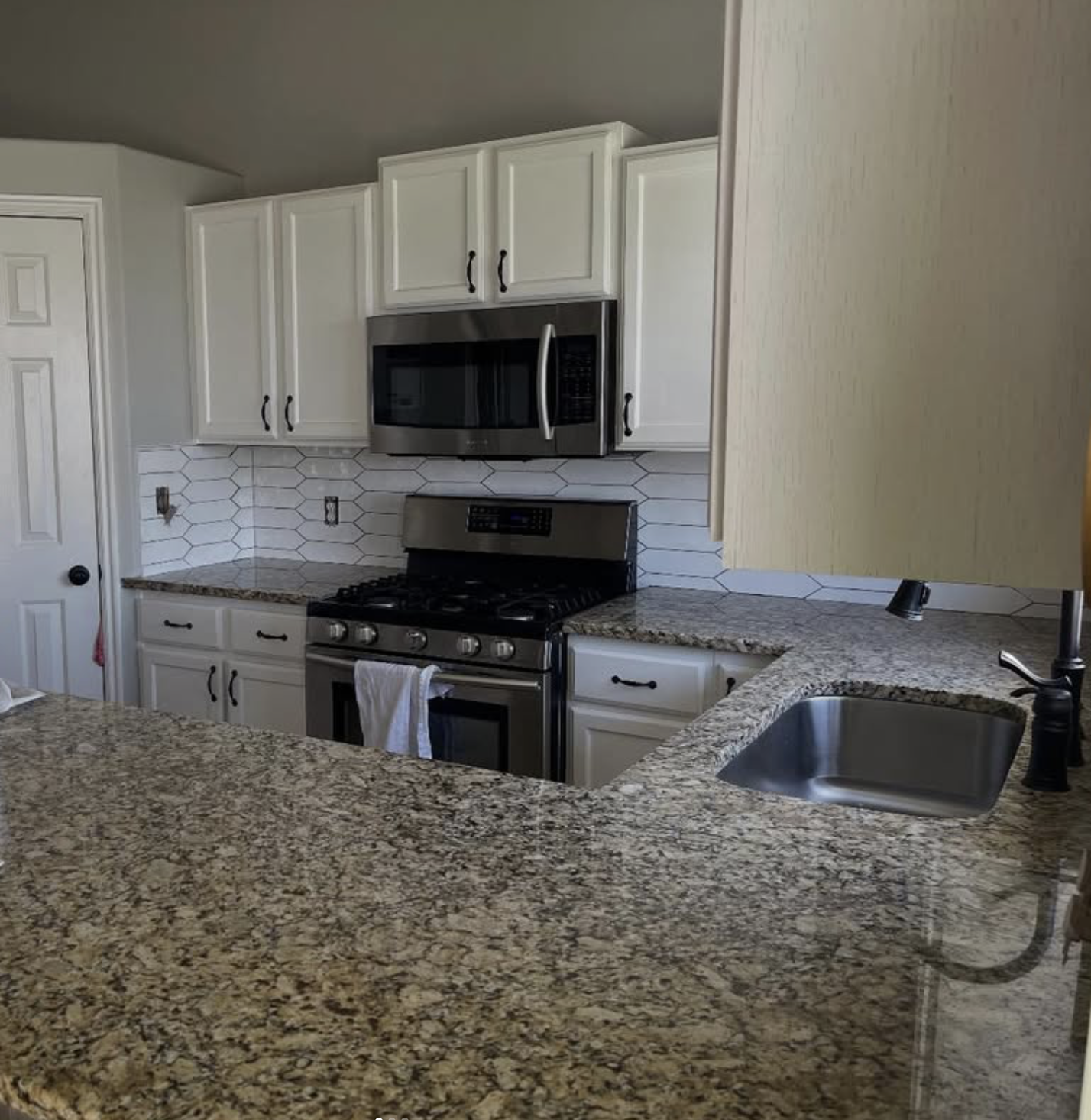 A kitchen with white cabinets, a granite countertop, a stainless steel microwave, a stove, and a small sink with a black faucet.