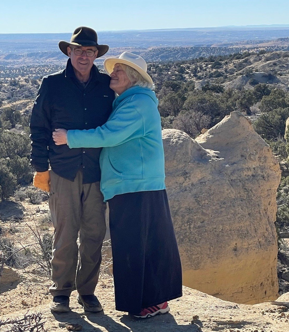 Two older women wearing hats hugging on a rocky outdoor landscape with hills and sparse vegetation in the background.
