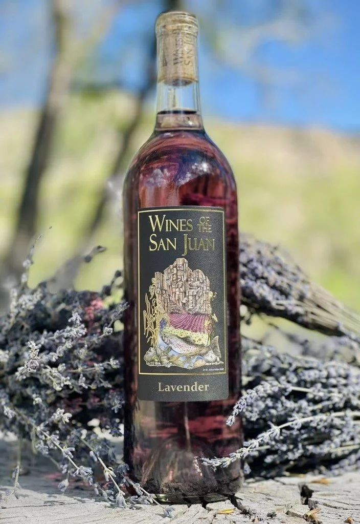 A bottle of lavender-flavored wine from Wines of San Juan is placed outdoors on a wooden surface surrounded by dried lavender sprigs. The background is blurred with a hint of trees and blue sky.