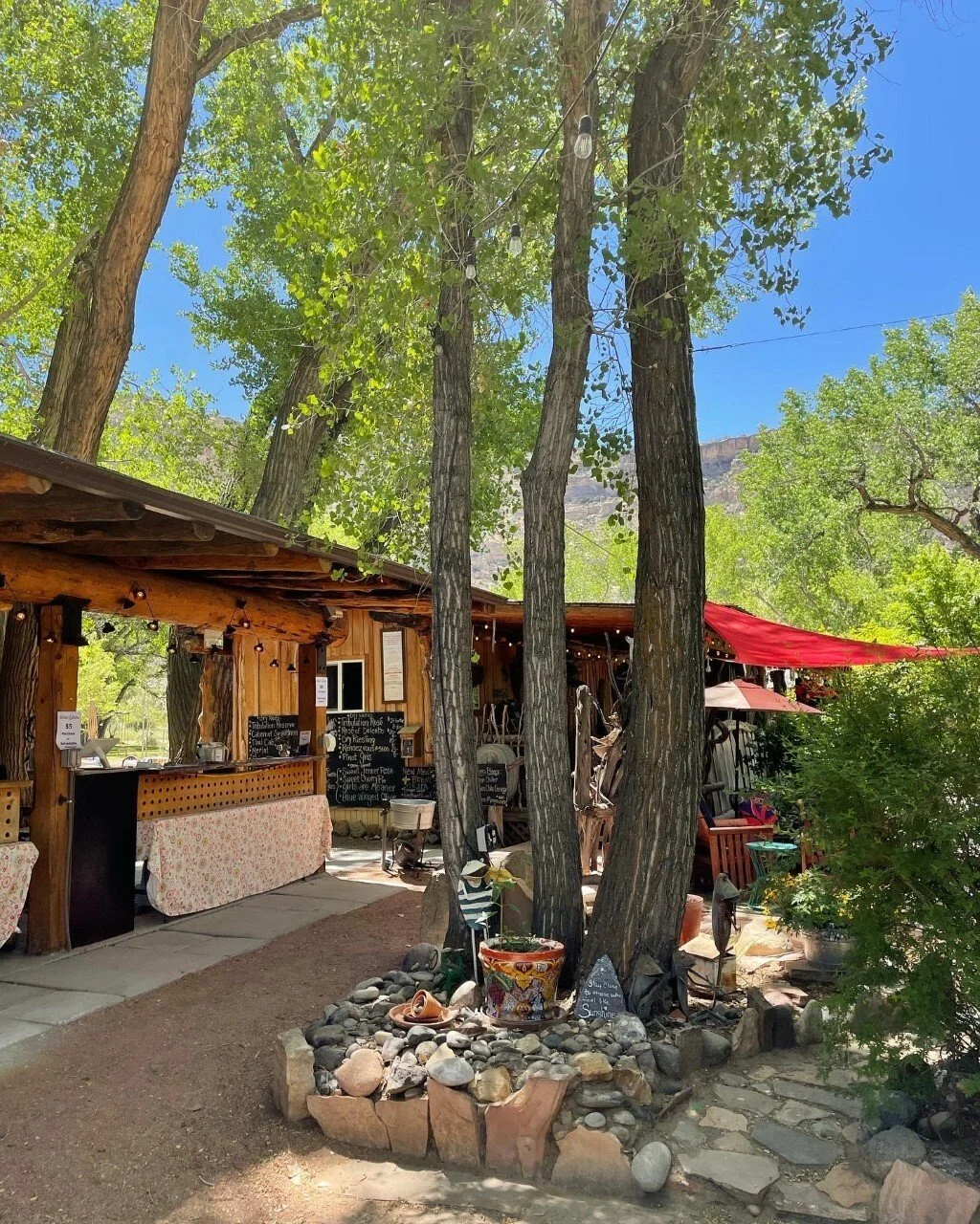Outdoor courtyard at Wines of the San Juan, rustic wooden food stand or shop with string lights, surrounded by trees, stone pathway, and outdoor seating with umbrellas.