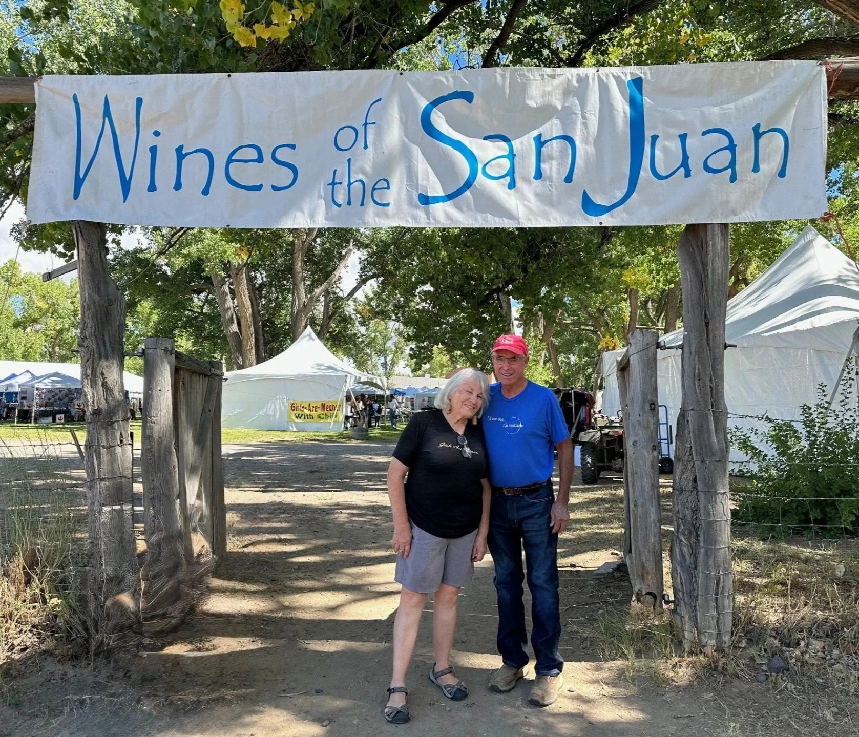 Two people standing under a sign that reads 'Wines of the San Juan' at an outdoor event, with white tents and trees in the background.