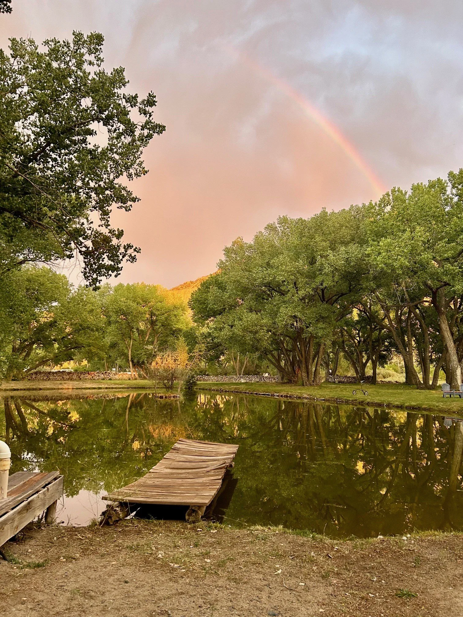 A peaceful scene of a small pond with an old wooden dock, surrounded by lush green trees, with a faint rainbow in a cloudy sky and reflections on the water.