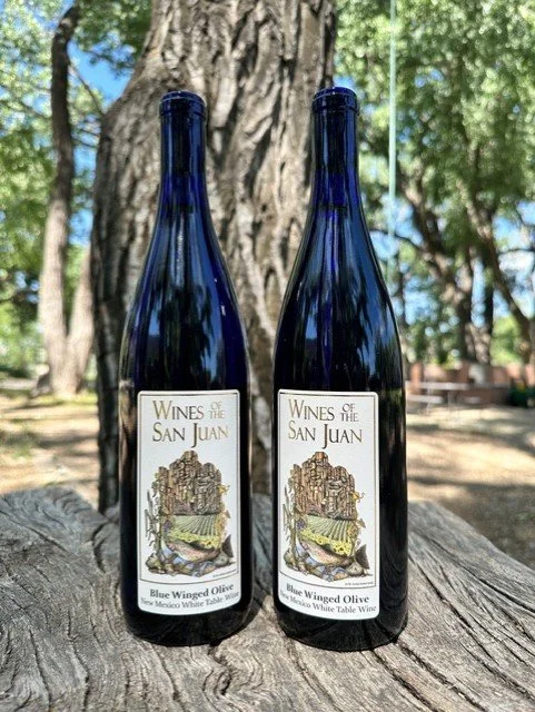 Two dark blue wine bottles labeled "Wines of the San Juan" and "Blue Winged Olive" standing on a wooden surface outdoors with trees and a blue sky in the background.