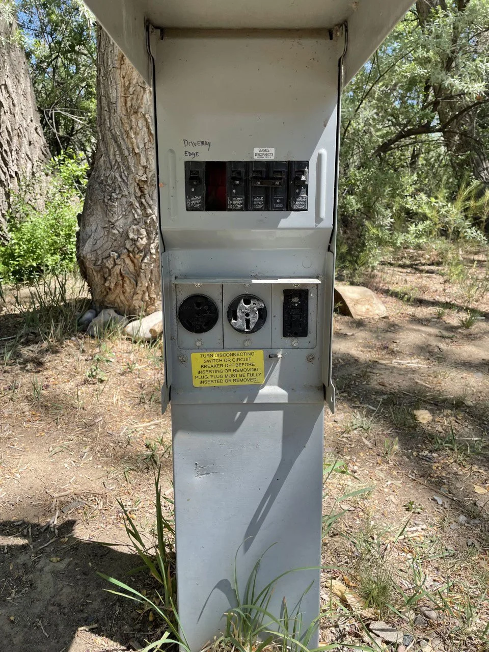 A gray outdoor electrical panel with circuit breakers, power outlets, and a warning label, situated near trees and grass.