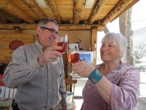 A man and woman clinking glasses with pink drinks, smiling at each other inside a rustic wooden structure.