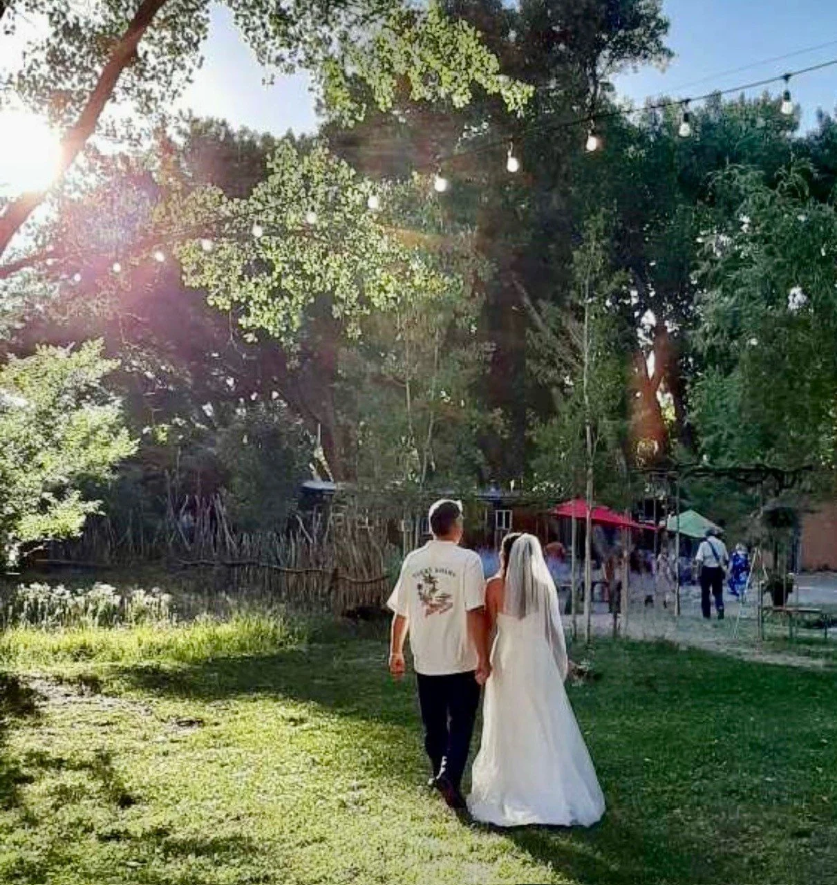 A couple, just married at Wines of the San Jua with the woman in a white wedding dress and the man in casual clothes, walking hand in hand through a park-like setting with green grass and trees, during the day with sunlight shining through the trees.