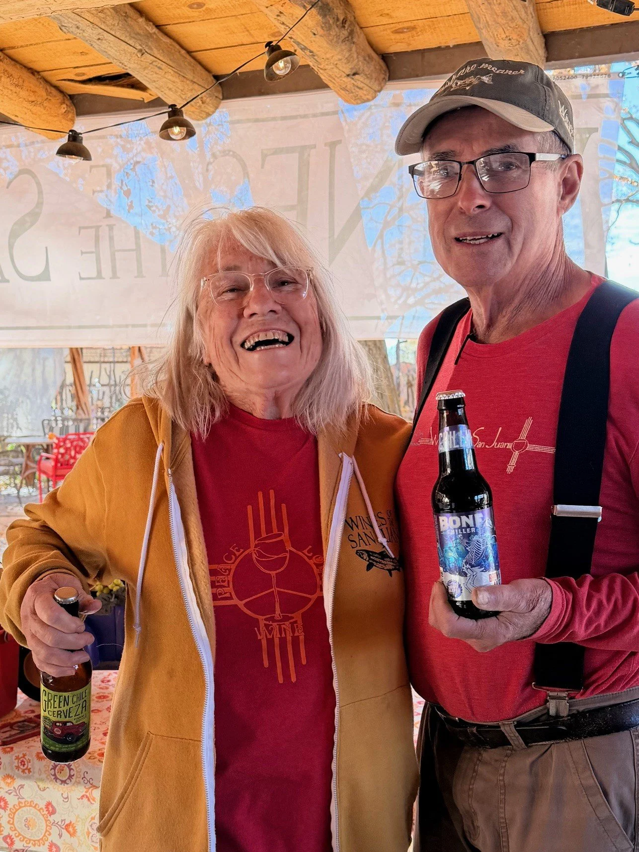 Two elderly people, a woman with blonde hair and glasses, and a man with glasses and a cap, are smiling and standing close together outside under a wooden roof. They are holding bottled drinks and appear to be at a social gathering.