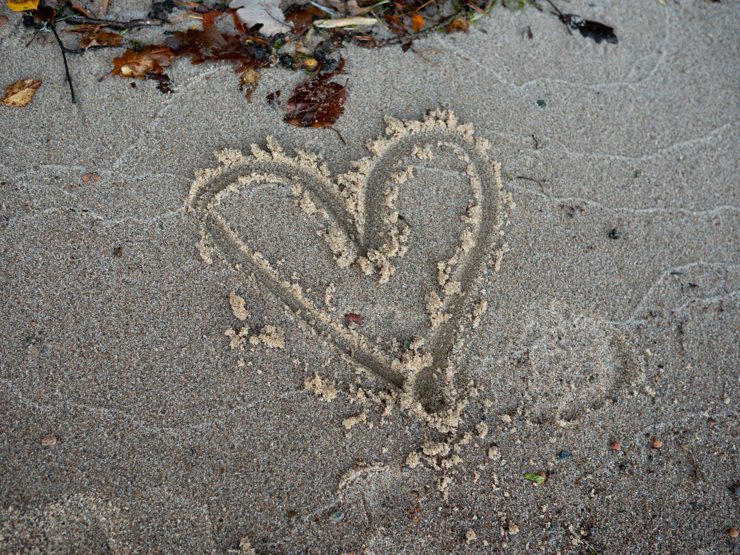 A heart drawn in the sand with a ring on the left side of the heart and some scattered leaves nearby.