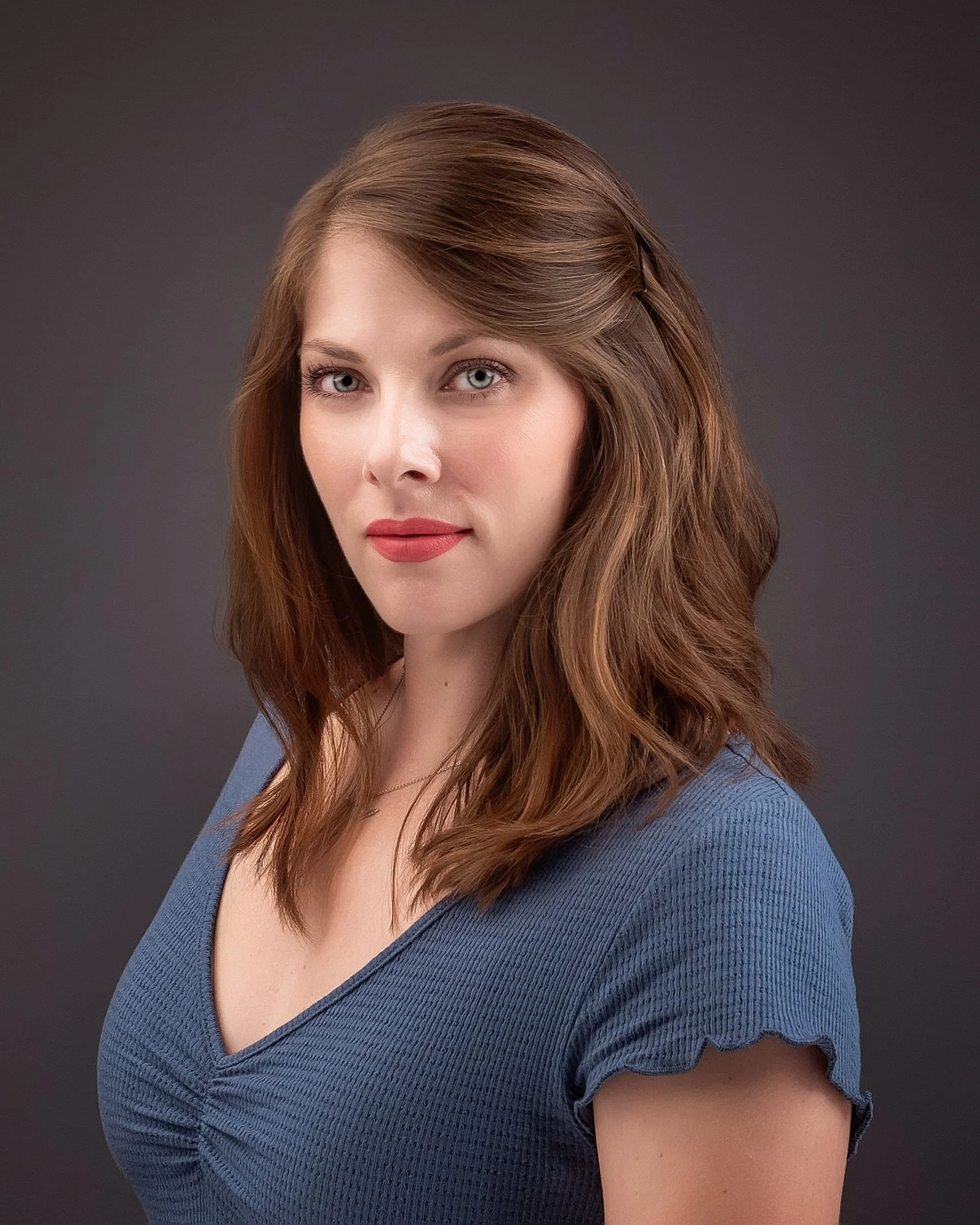 Portrait of a woman with light skin and wavy brown hair, wearing a blue top, against a dark background.