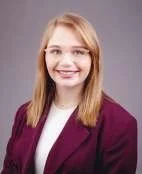 Portrait of a young woman with blonde hair, smiling, wearing a maroon blazer over a white top.