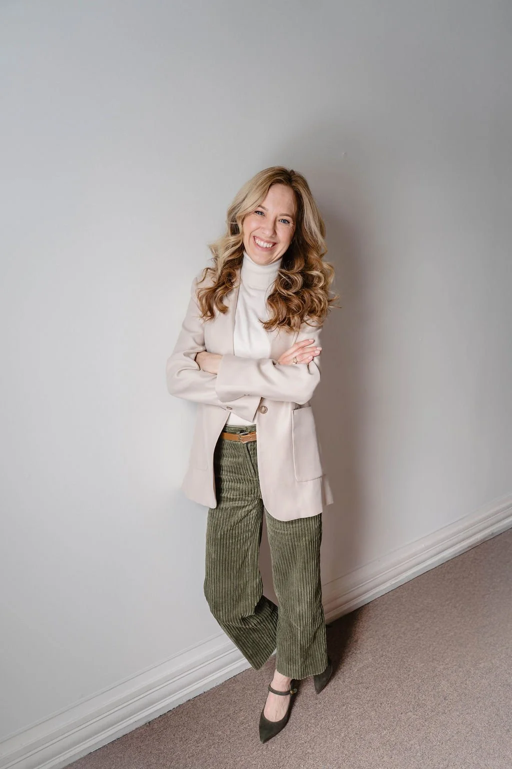 A woman with blonde curly hair standing against a plain white wall, smiling, wearing a beige blazer, white turtleneck, olive green corduroy pants, and black shoes.