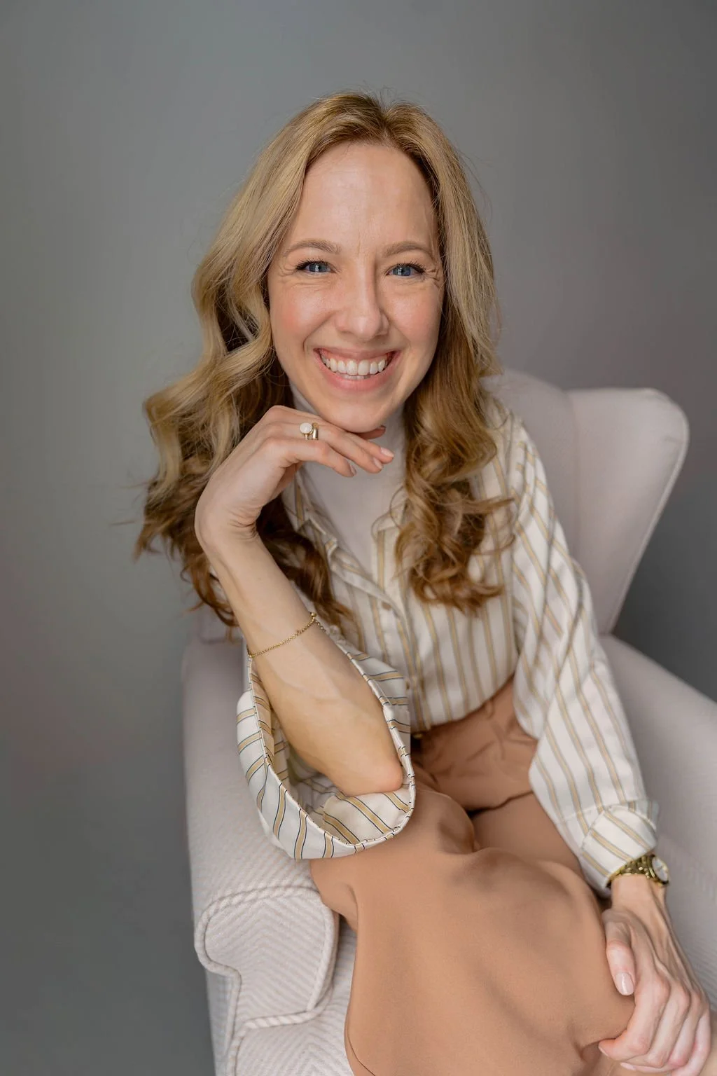 A woman with curly blonde hair smiling, sitting on a white chair, wearing a striped blouse and beige pants, with her hand resting on her chin.