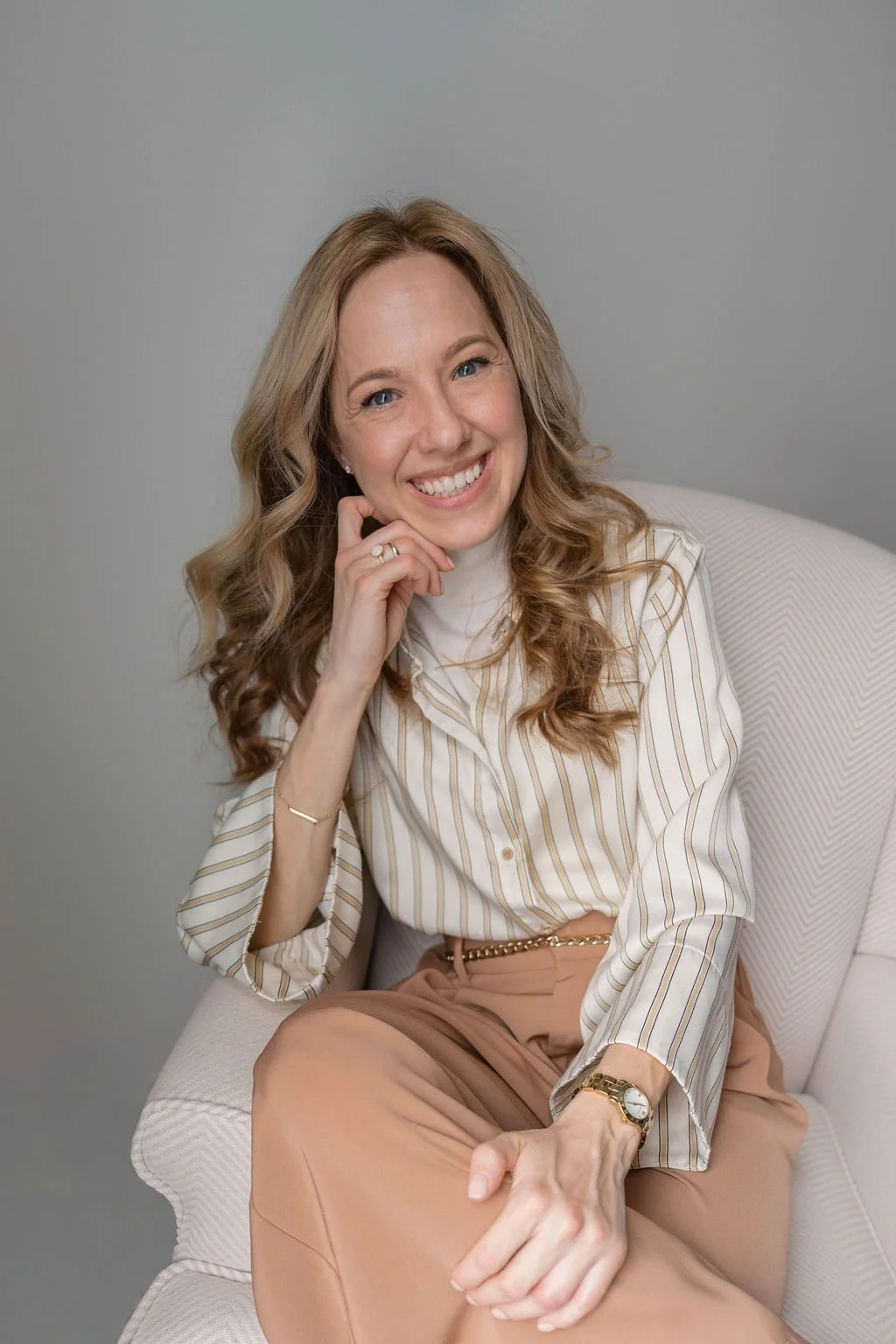 A woman with long, wavy blonde hair, smiling and sitting on a light-colored chair against a plain gray background. She is wearing a cream and gold striped blouse, beige pants, and accessorized with a watch, a bracelet, rings, and a chain belt.