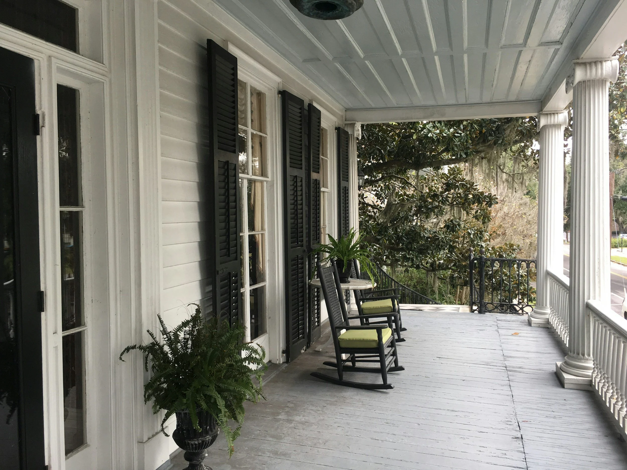 Front porch with white wood siding, black shutters, black rocking chairs with green cushions, potted plants, and tall white columns.