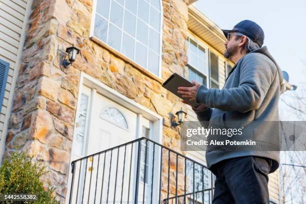 A man wearing a hoodie, sunglasses, and a cap holding a clipboard while standing outside a stone house with solar panels on the roof.