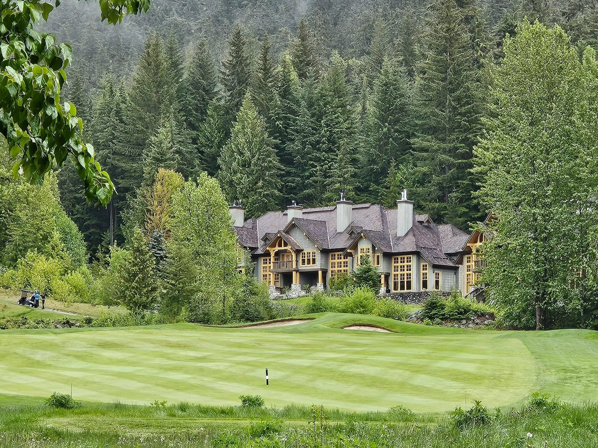 A golf course with green grass, sand traps, and a golf ball marker in the foreground. In the background, there is a large house with multiple chimneys, surrounded by tall trees and dense forest.