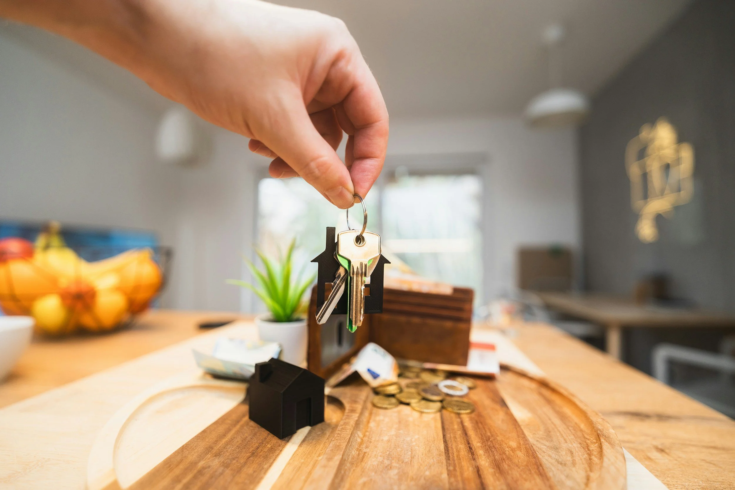 Hand holding a keyring with house-shaped keychains over a wooden table with coins, a small house model, and a plant in the background.