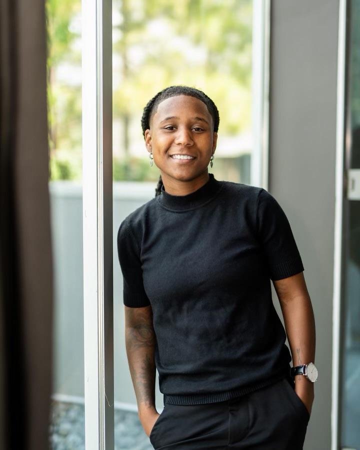 A woman dressed in all black, with hands in her pocket leaning before a glass door.