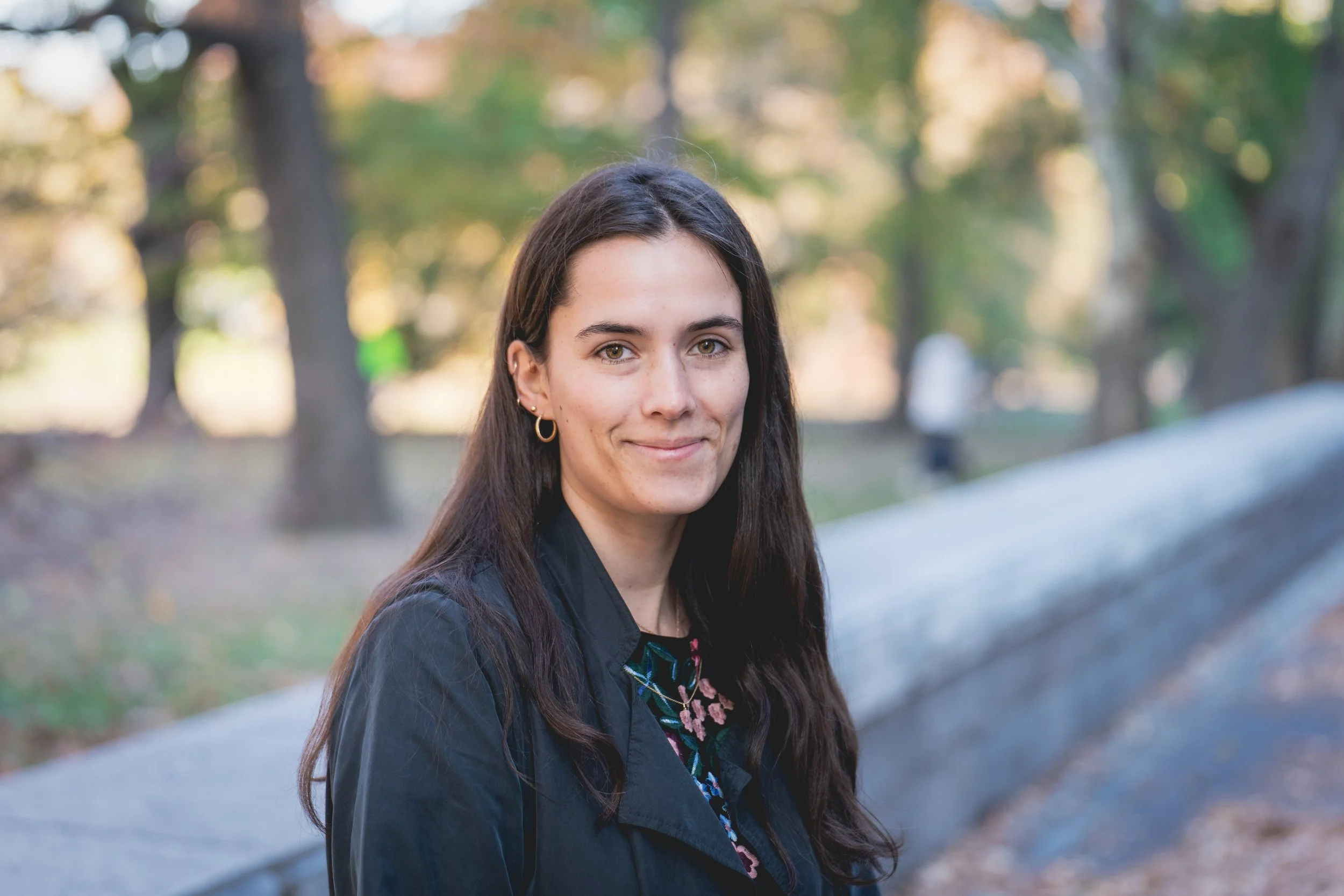 A woman with long dark hair and earrings smiling outdoors in a park with trees in the background.