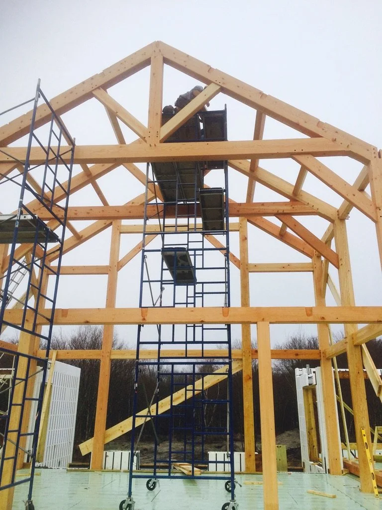 Construction of a wooden house framing with scaffolding and a worker at the top, against an overcast sky.