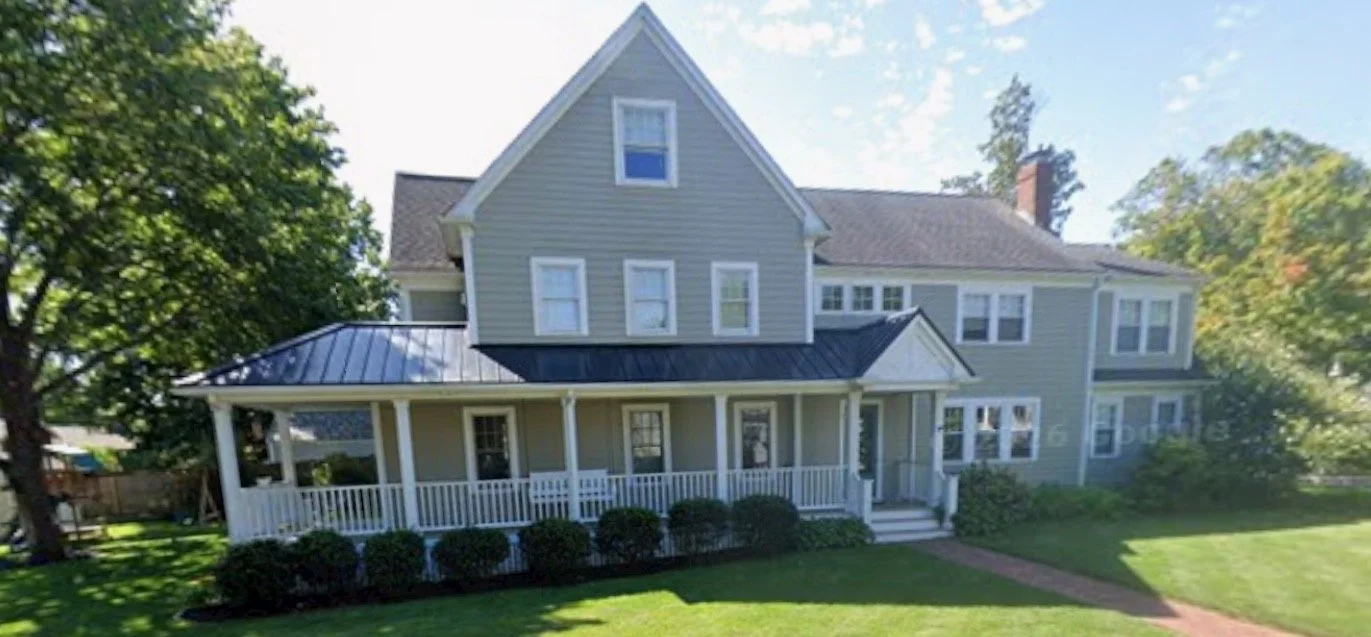 A large two-story house painted light grey with white trim, featuring a front porch with white railings, multiple windows, and a metal roof on the porch and part of the house. The house has a lawn with bushes and trees surrounding it.