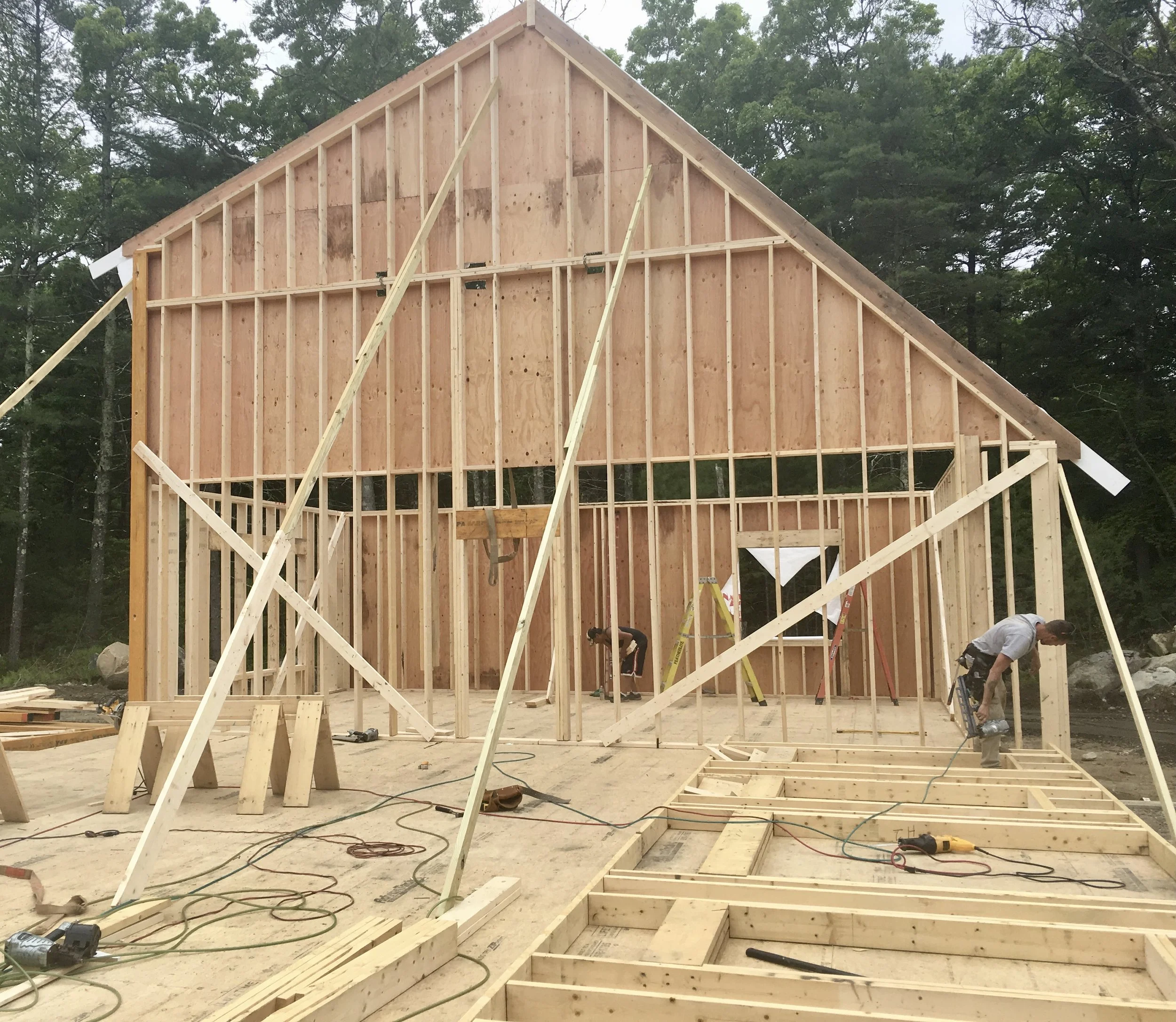 Construction of a wooden house frame in a forested area, with workers installing the structure while surrounded by construction tools and materials.