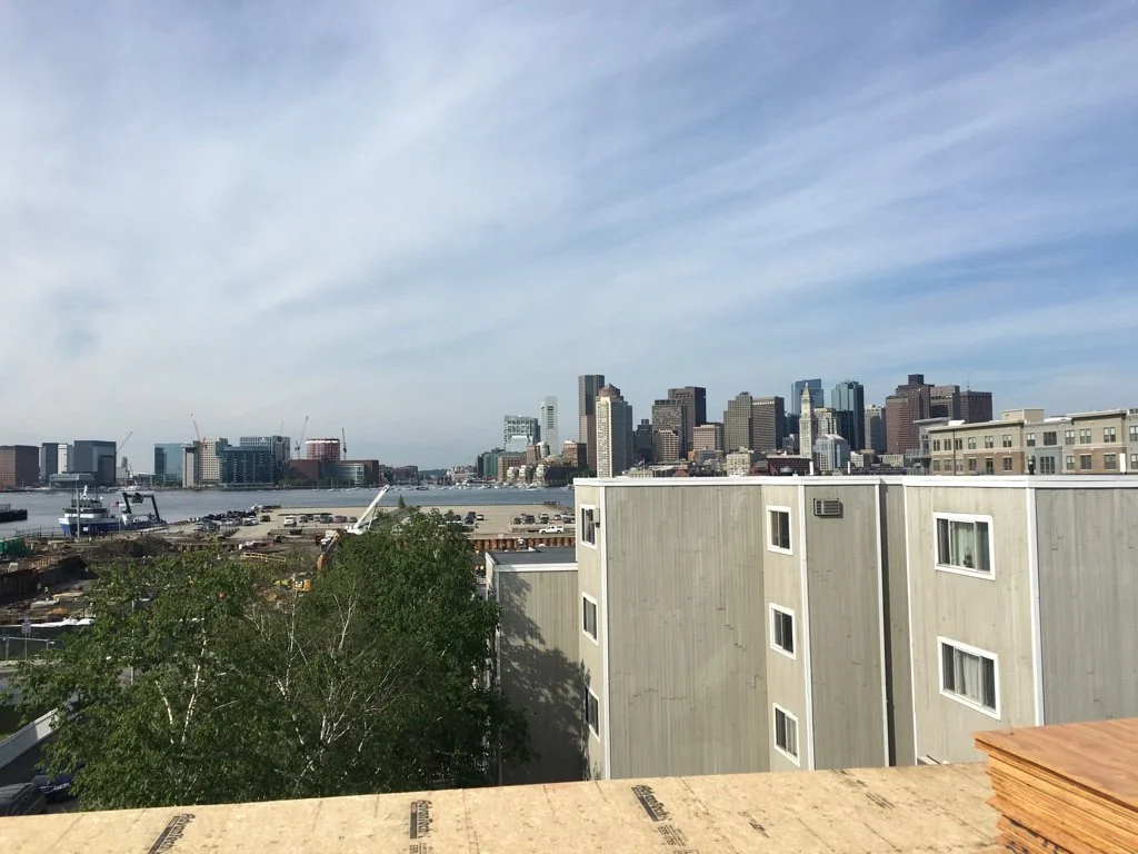 City skyline with tall buildings across a body of water, with a partly cloudy sky and some construction cranes visible. In the foreground, there are beige residential buildings, some trees, and construction materials.