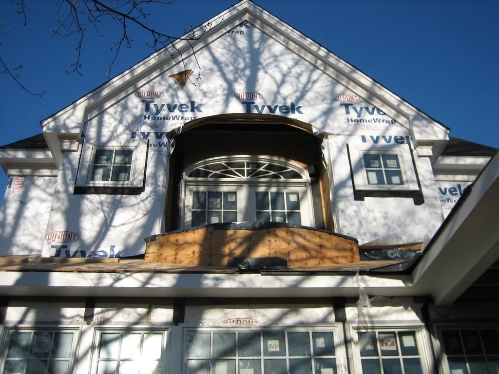 Front of a house under construction with Tyvek HomeWrap, three windows on upper floor, decorative window with arch and small panes in the center, and a wooden porch or balcony area.