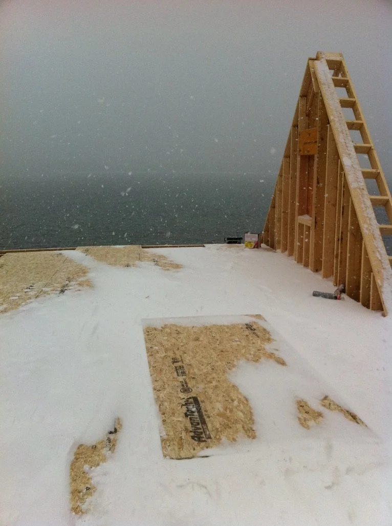 Construction site on a snowy day with a wooden frame structure and plywood sheets, overlooking an ocean in the background.