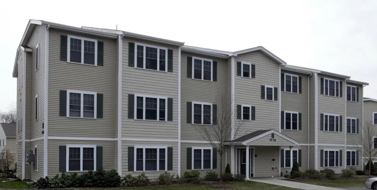 A three-story residential apartment building with beige siding and white trim, featuring multiple evenly spaced black-shuttered windows and a small entry porch.
