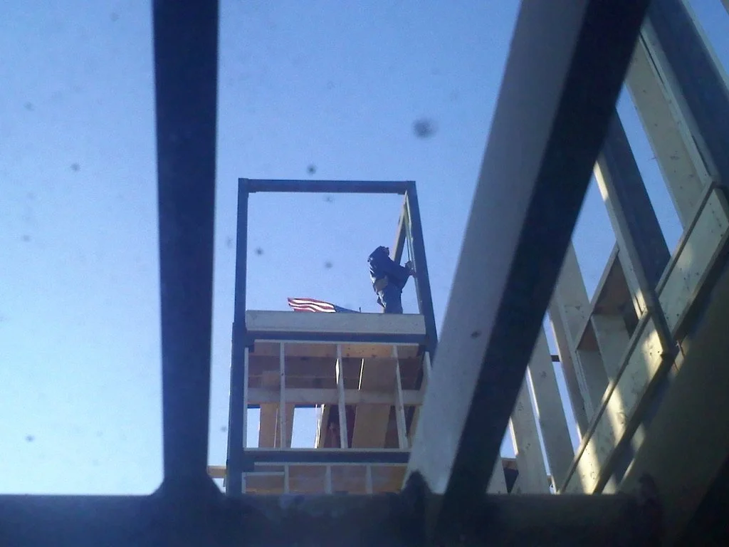 View of a construction worker on a building's scaffold, seen from below through a metal frame, with a clear blue sky in the background.
