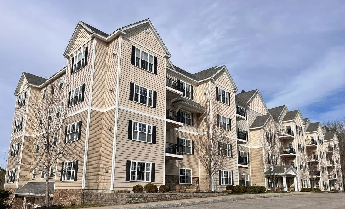 Multi-story apartment building with beige siding, black shutters, and balconies under a cloudy sky.